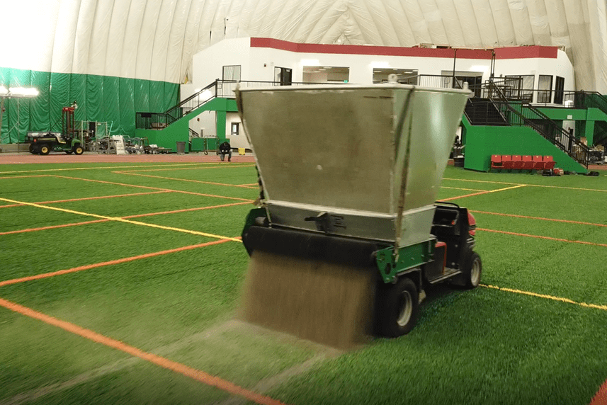 A machine fitted with a hopper spreads sand over the top of a turf field.