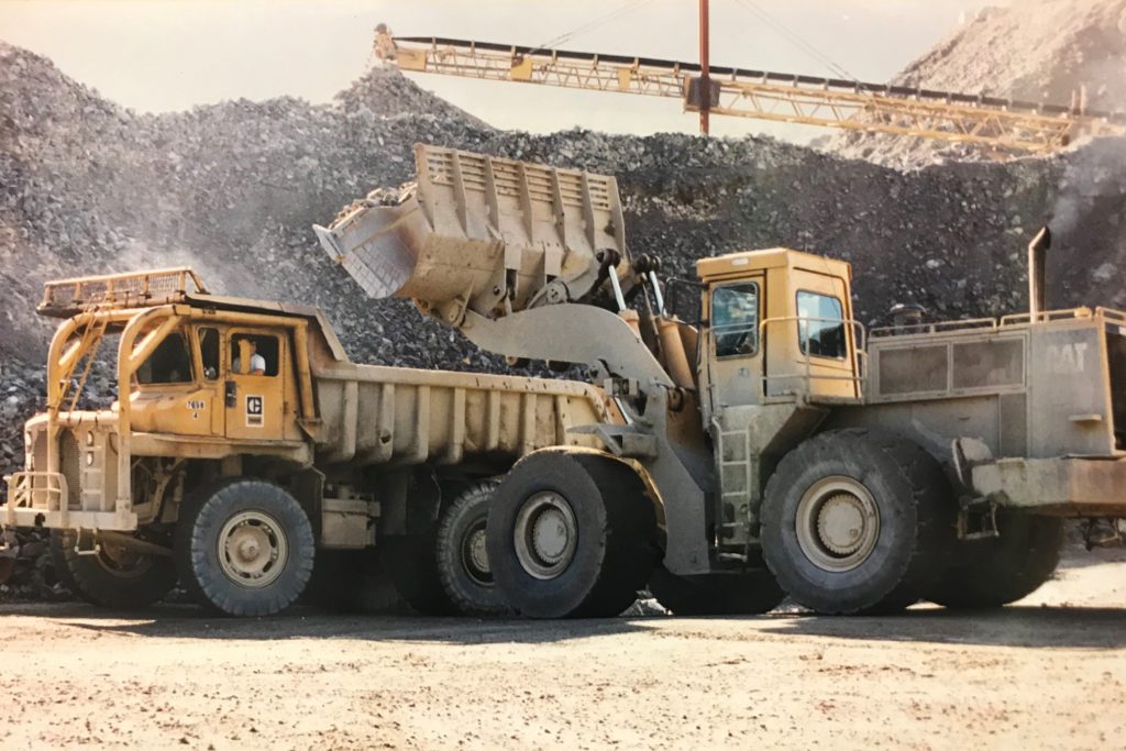 A loader machine prepares to pour rock into a large dump truck. A large pile of rock and a belt can be seen in the background.