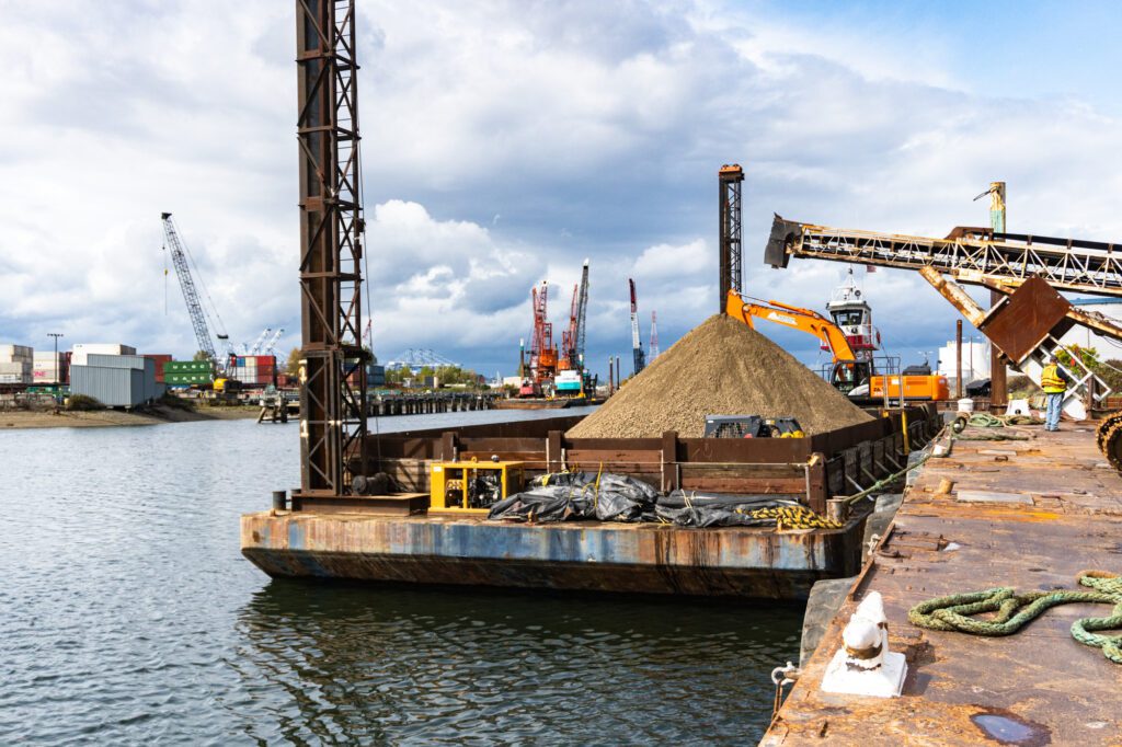 A barge filled with a rock-and-sand material floats in a waterway. Cranes can be seen in the background.