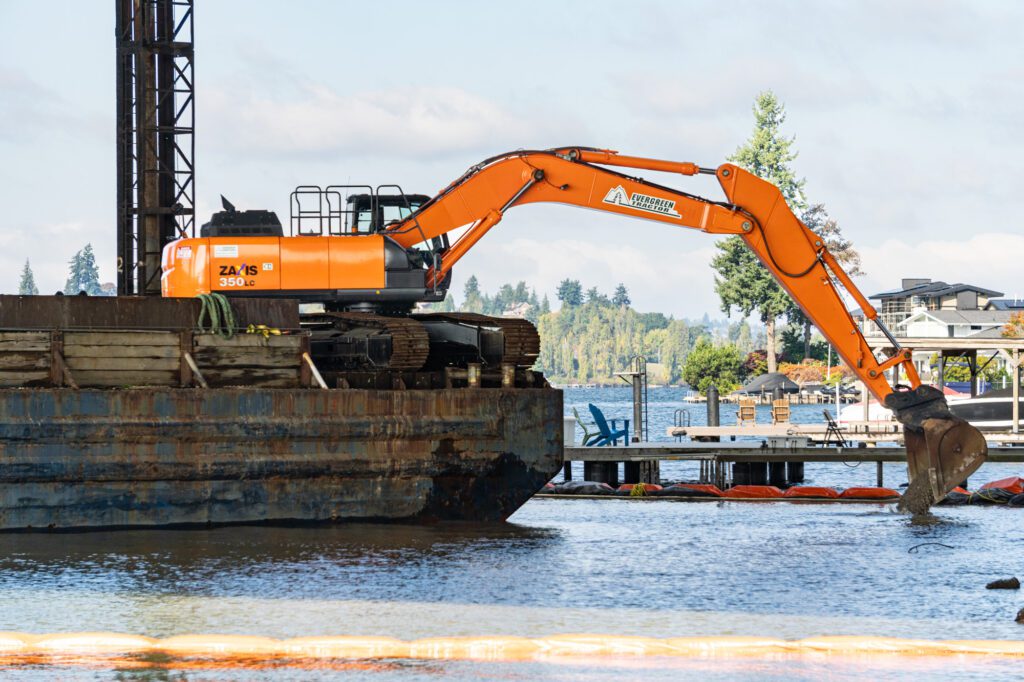 An orange excavator working at the edge of a barge pours a rock mixture into water. Docks and a large house can be seen in the background.