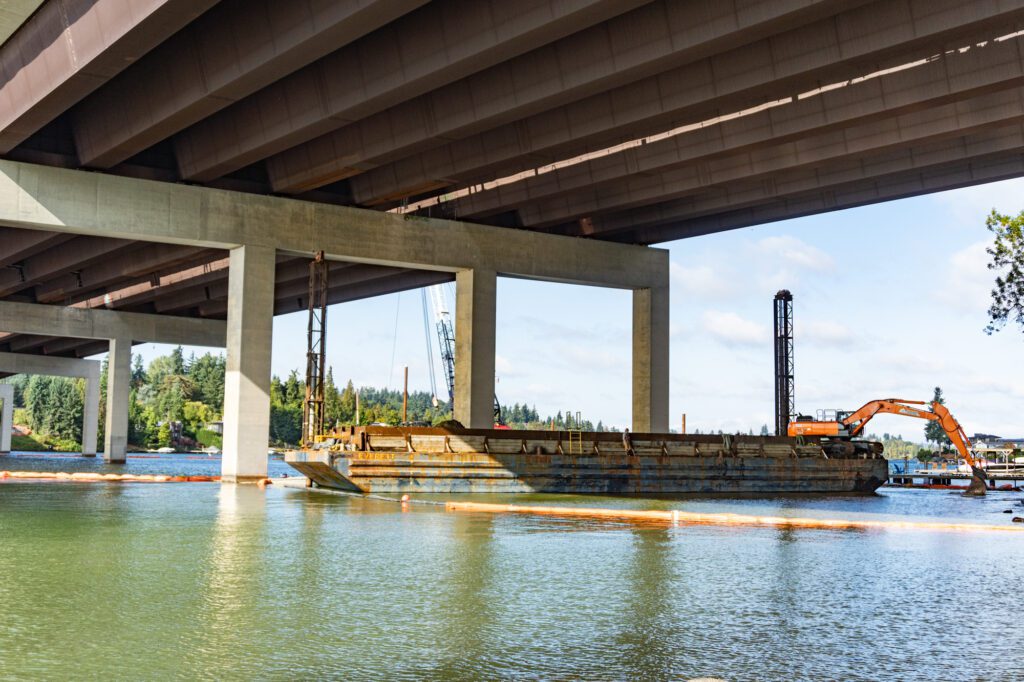 An orange excavator works at the end of a barge with its bucket reaching into the water. The barge floats underneath a highway overpass. The beams of the overpass can be seen at the top of the photo.