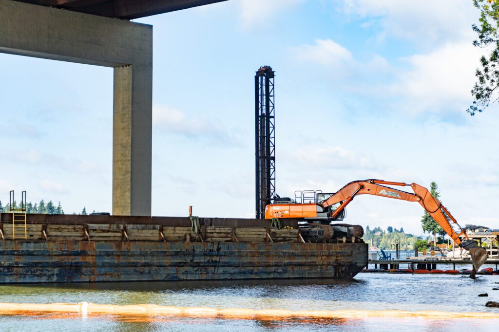 An orange excavator working at the edge of a barge pours a rock mixture into water. Docks and a large house can be seen in the background. The barge floats next to the columns of a highway overpass.