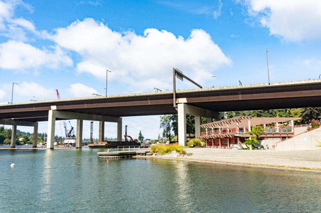 In the background, a barge works on the water under a highway overpass. A building with red trim is in the foreground next to a beach.