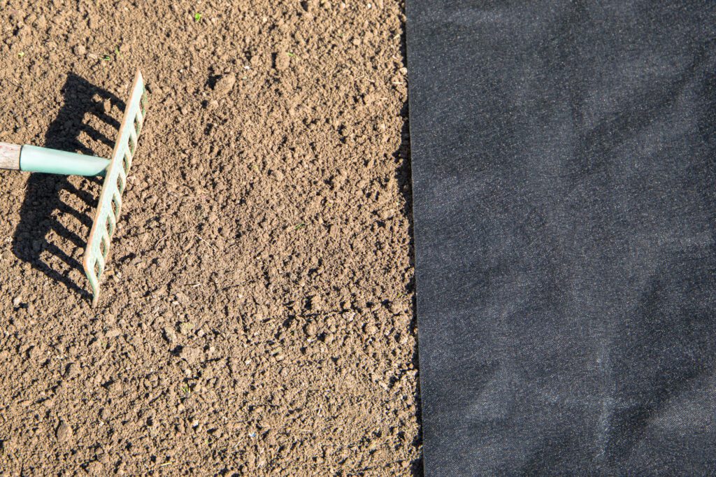 a piece of nonwoven landscape fabric sits on a prepared bed of soil where a garden rake rests