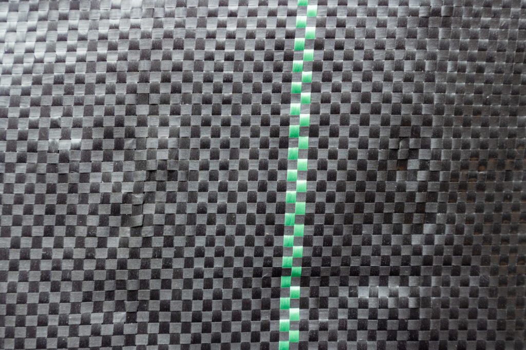 A closeup of a piece of woven landscape fabric shows how black threads crisscross like a woven basket. A green line runs down the middle of the fabric.