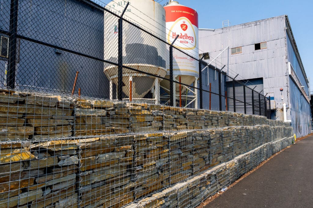 Two beer tanks sit above a tiered gabion wall that stops at the edge of a blue warehouse. The gabion cages are filled with Gold Rush quartzite, a type of rust-colored ledge rock.