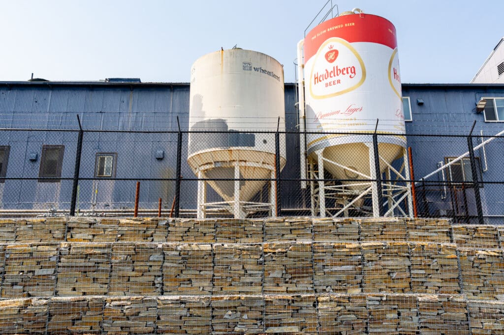 Two beer tanks sit above a tiered gabion wall. The gabion cages are filled with Gold Rush quartzite, a type of rust-colored ledge rock.