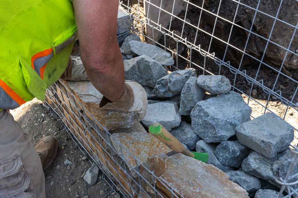the hands of a construction worker rest on the edge of an open gabion basket filled with rock