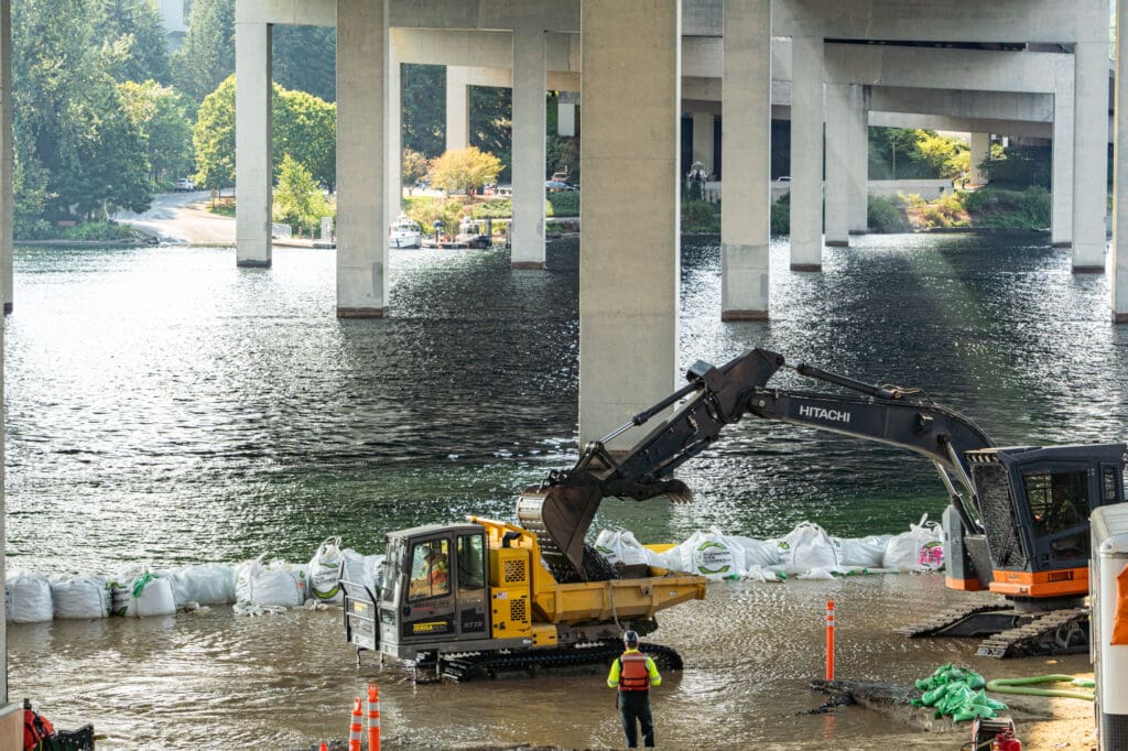 an excavator sits in the water along a shoreline and loads a crawler with rock for removal