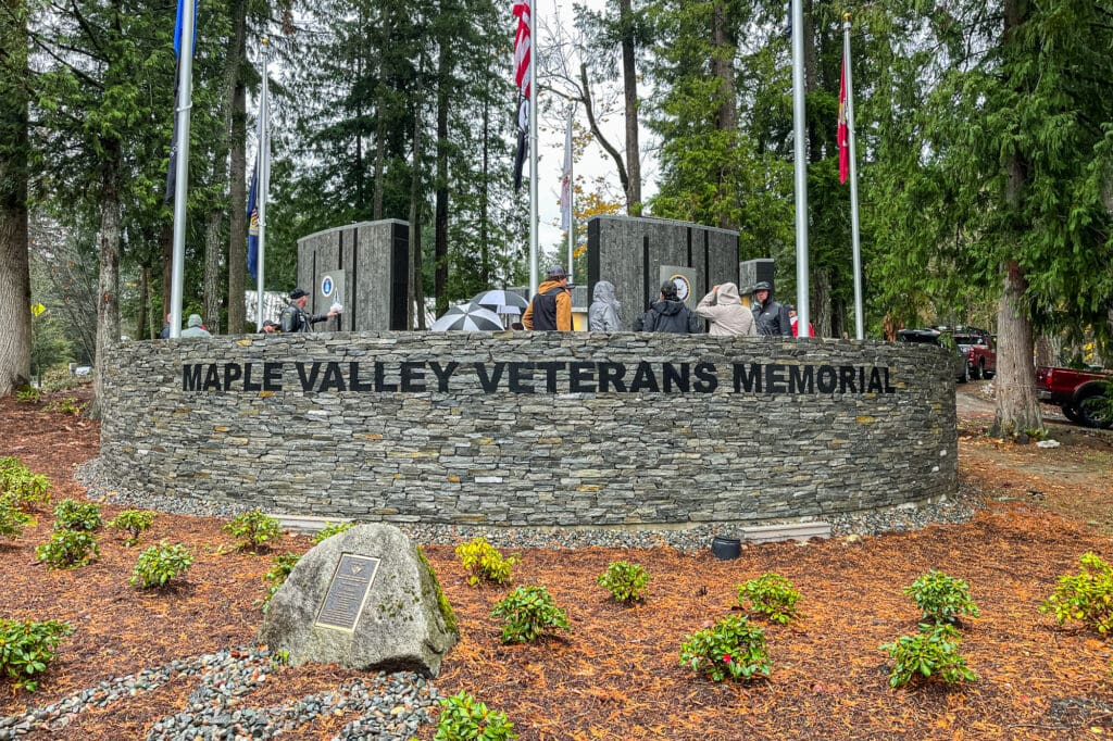 A memorial filled with large walls sits within a retention wall made up of flat stones. People stop at each wall, which represents a branch of the military, and look at the military plaques.
