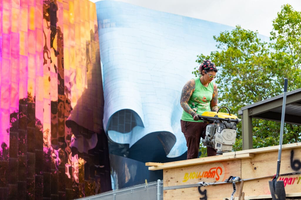An artist named Jamie Louks uses a plate compactor to compact sand inside a form. An iridescent purple glass building and a sky blue building can be seen in the background.
