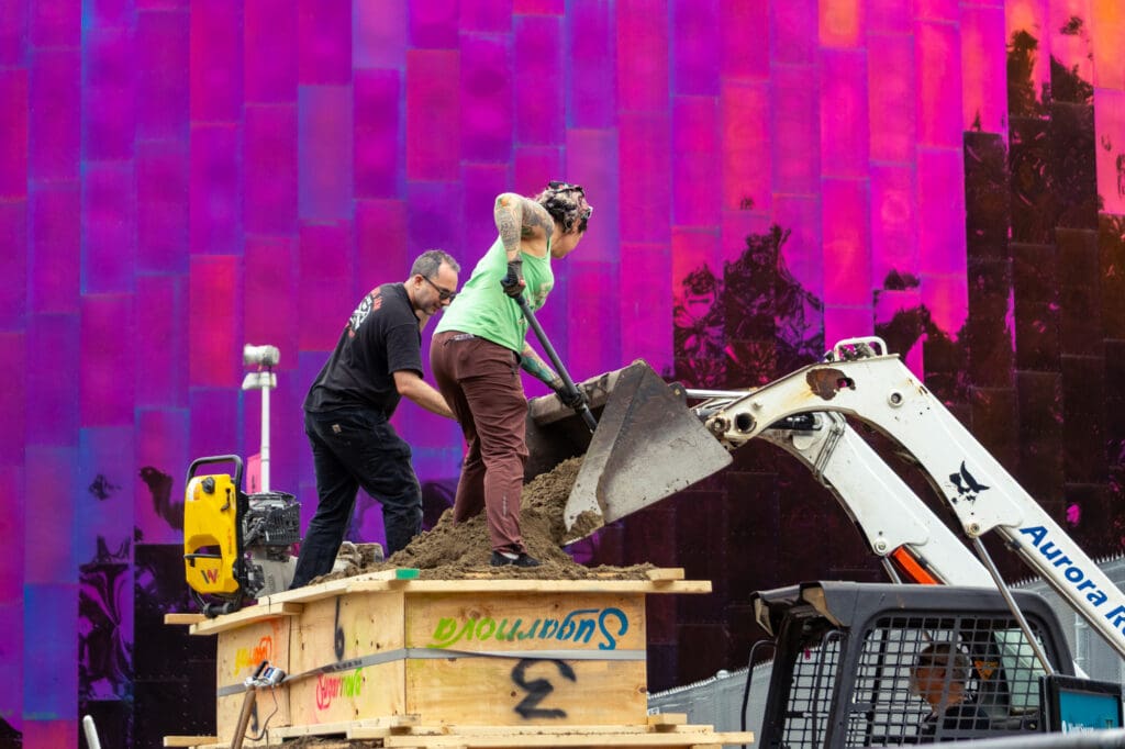 Two people use shovels to unload the bucket of a skidsteer into a form for a sand sculpture.