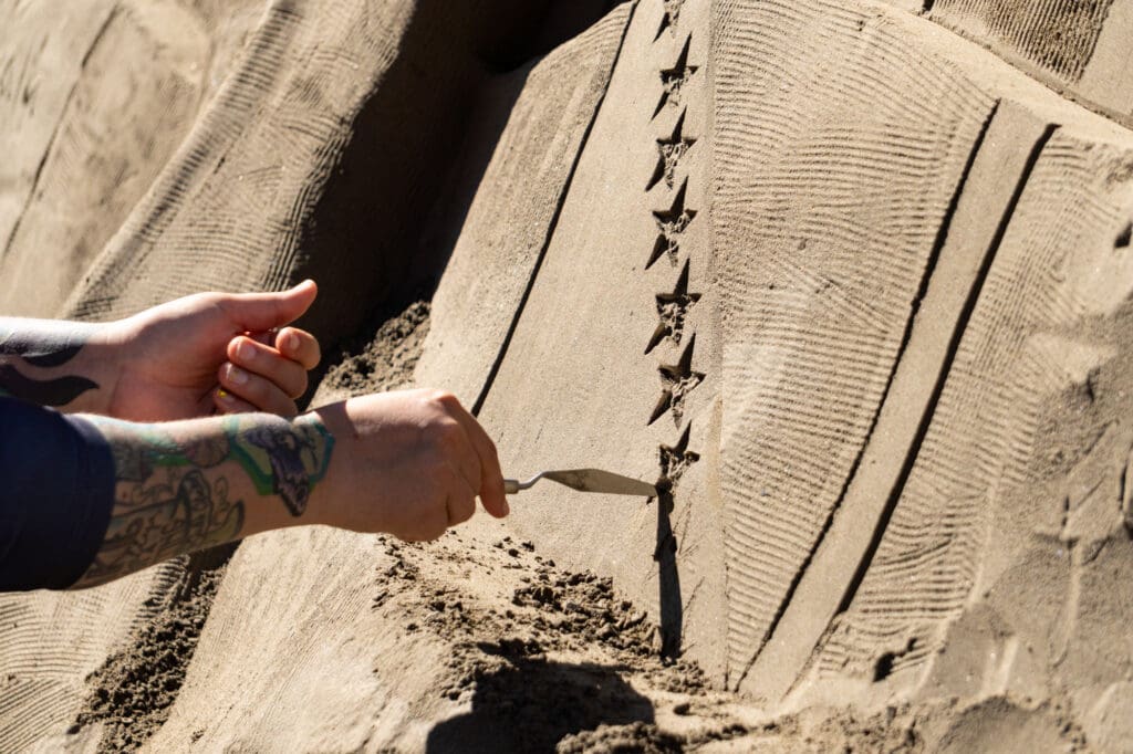 a closeup of an artist's hand holding a palette knife and carving stars into a sand sculpture