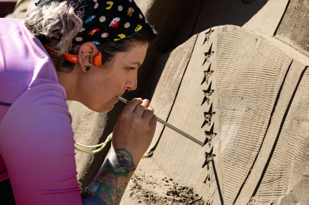 an artist blows into a metal straw to blow sand away from details on a sand carving