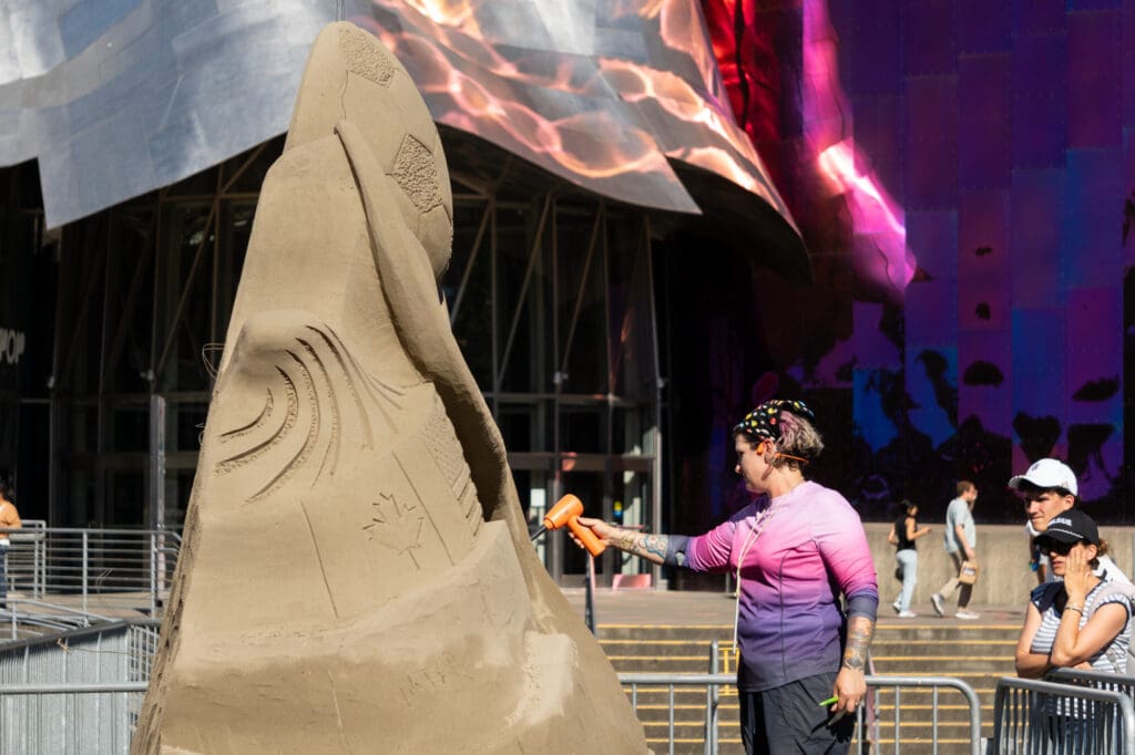 an artist uses an air duster to blow loose sand off of a tall sand sculpture