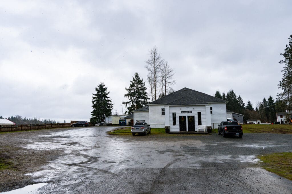 Light reflects off the slick surface of a scrappy gravel driveway. The scene is overcast. A white country-style building can be seen in the background.
