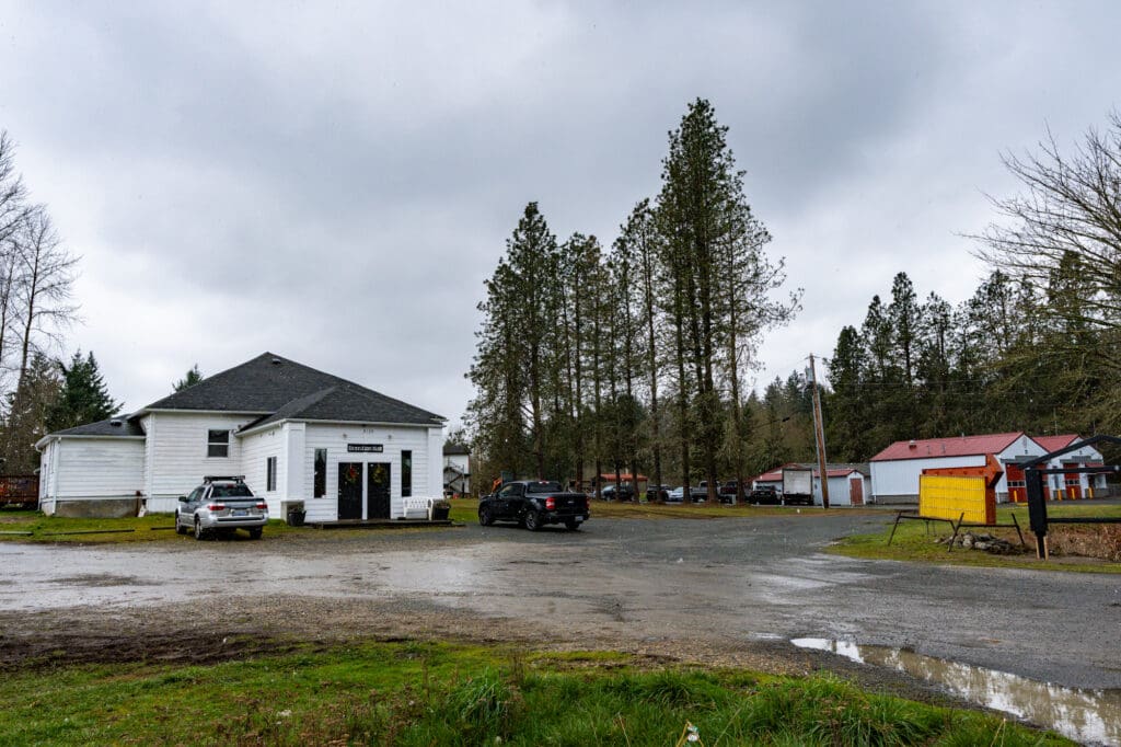 A wet, scrappy gravel driveway, flanked by a large puddle, leads to a white country-style building. The scene is overcast.