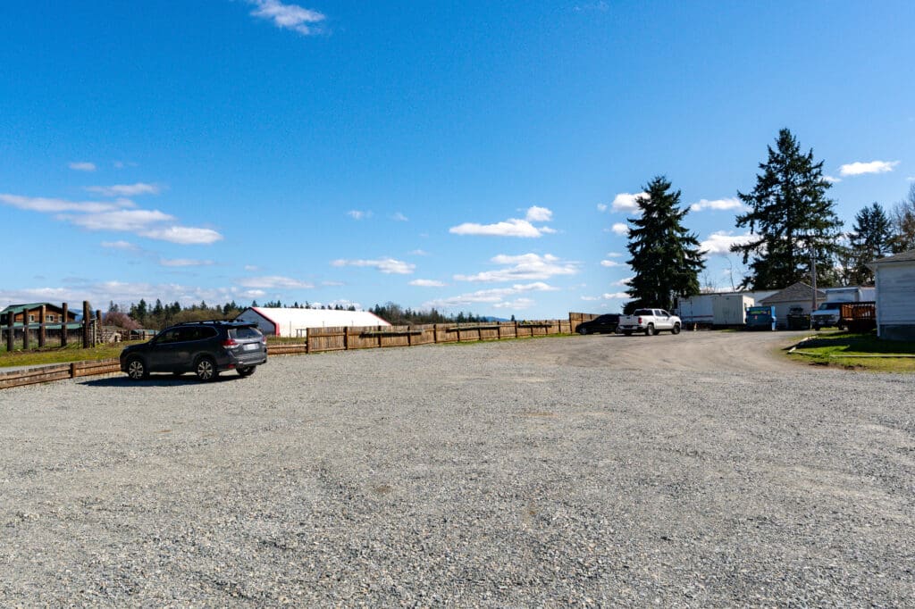 A broad gravel driveway winds past a fence next to a ranch. The scene is sunny.