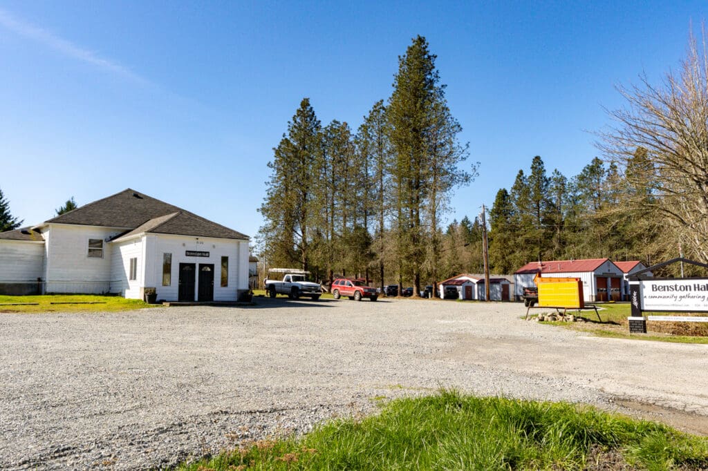 A gravel driveway, flanked by grass, leads to a white country-style building. The scene is sunny.