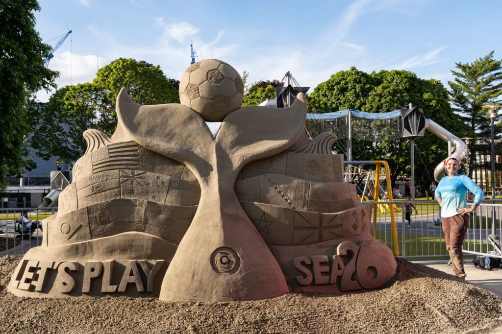 A woman stands next to a sand sculpture that is nearly double her height. The sculpture features a giant whale tail balancing a soccer ball, surrounded by 