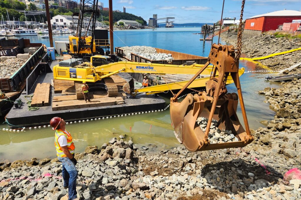 The rock-filled bucket of a crane, perched offshore on a barge, hovers over a rocky shoreline while a construction worker looks on.