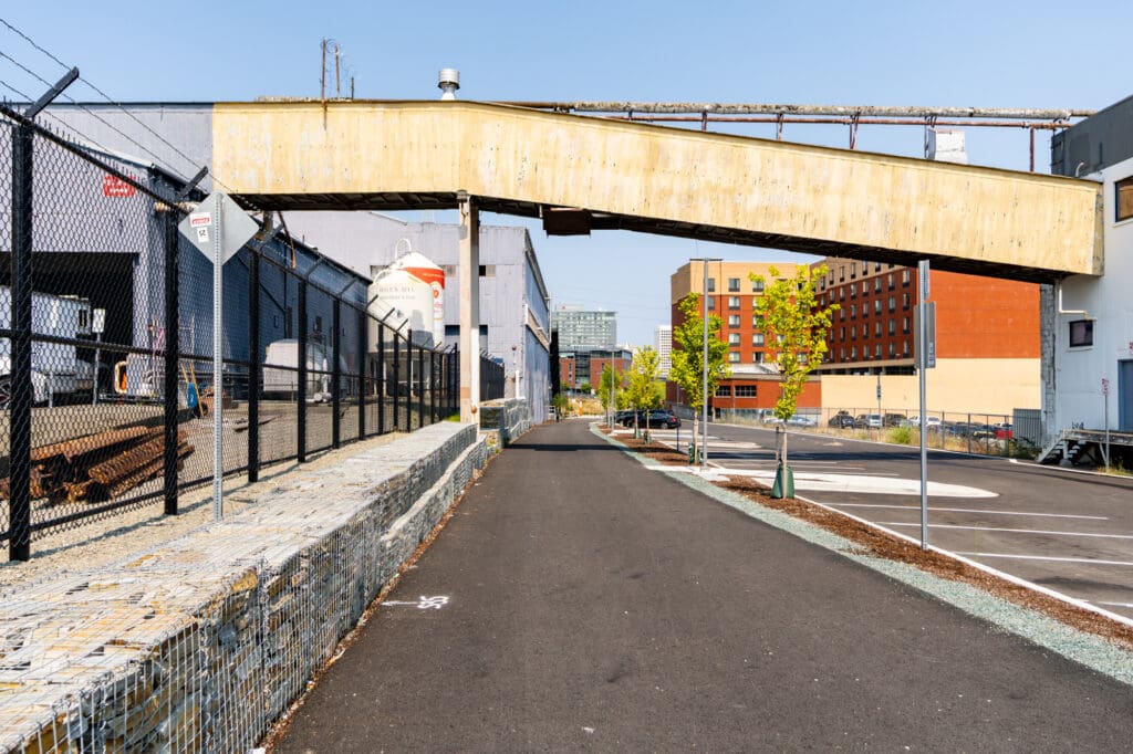 A paved path runs through an alley between warehouses, bordered by a landscaped parking lot and gabion walls filled with ledge rock. A skybridge-like structure adjoins two warehouses above the path.