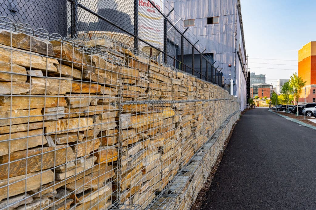 A gabion wall runs the length of a fence. The gabion cages are filled with Gold Rush quartzite, a type of rust-colored ledge rock.