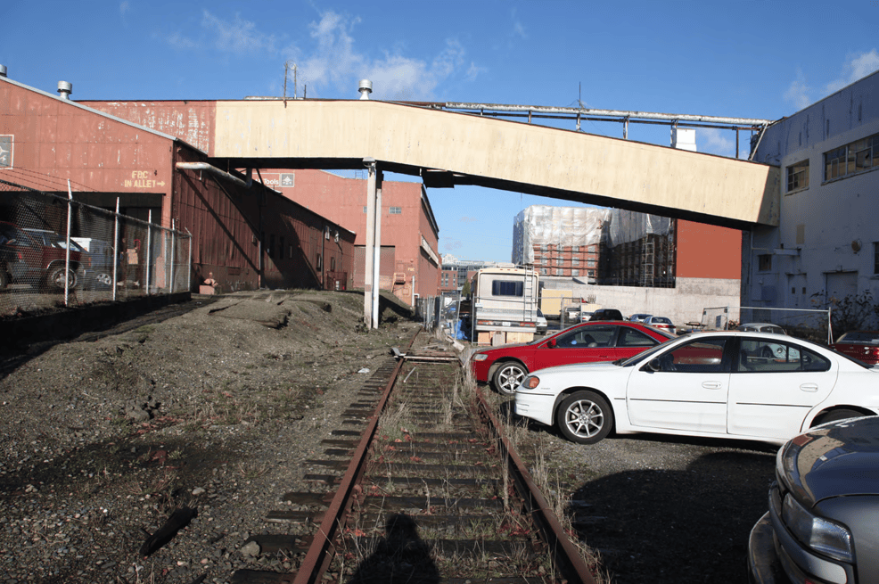 Railroad tracks, overgrown with weeds, run through an alley between warehouses. Cars are parked along the edge of the defunct track.