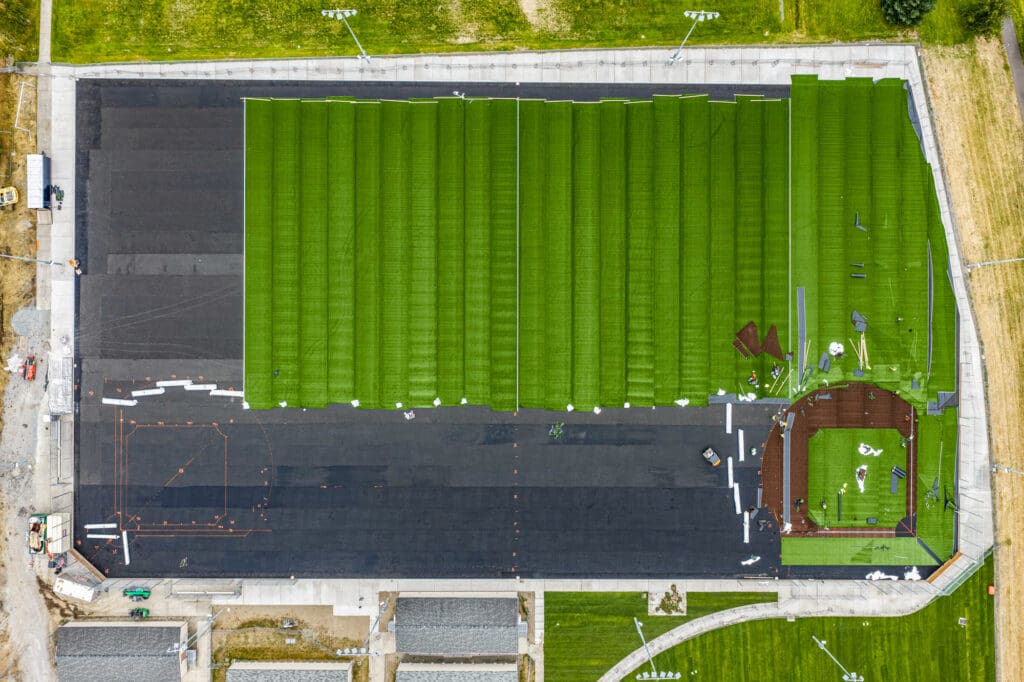 An aerial view shows strips of synthetic turf covering most of an E-Layer system (a pavement-like coating made up of epoxy and round stones).