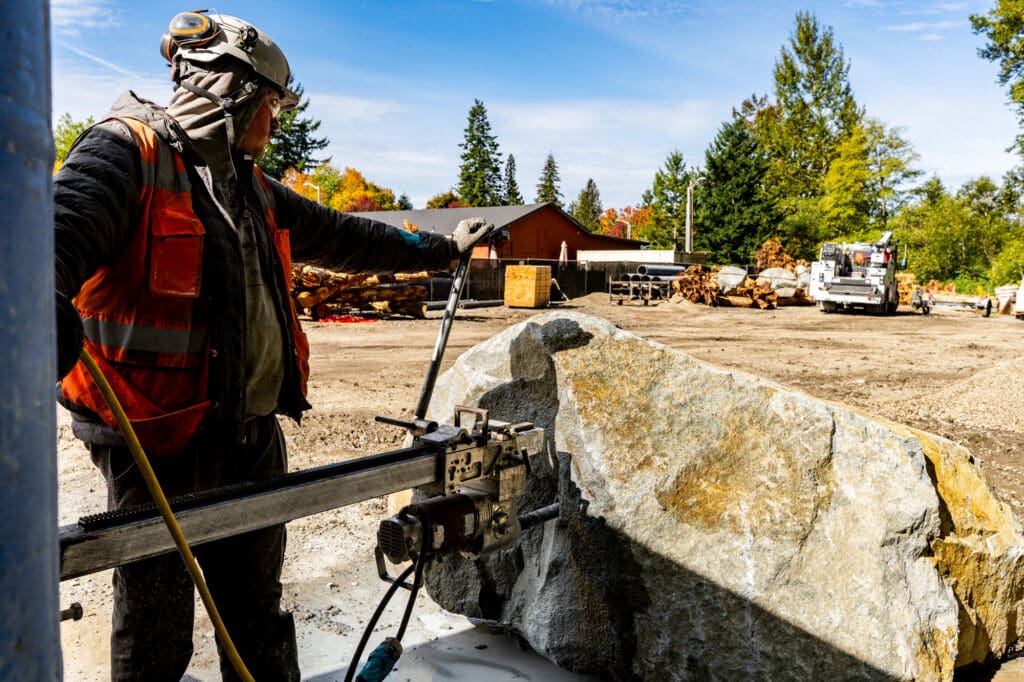 A worker in construction apparel operates a drill as it drills through an oversized piece of quarried rock.