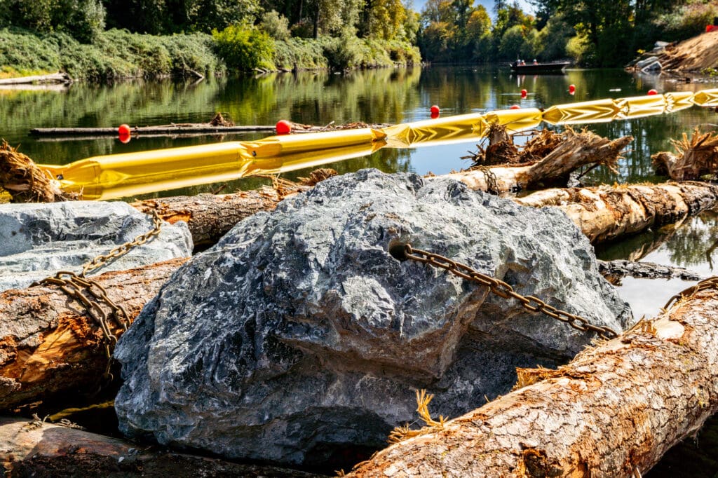 An enormous quarry boulder is anchored to a log in the river next to a riverbank.