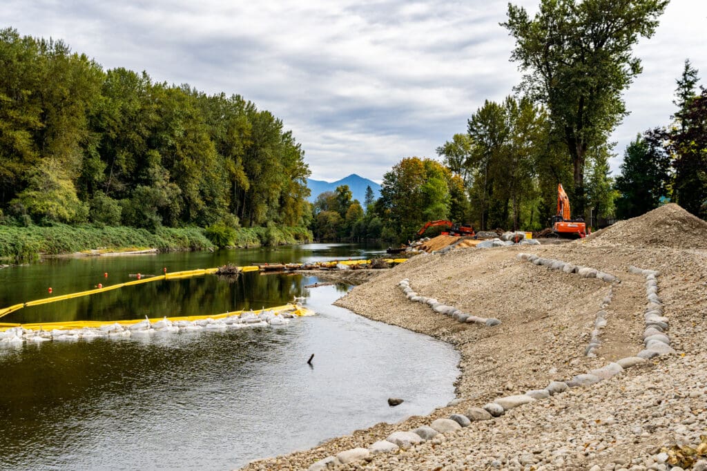 A rocky riverbank appears to be under construction. The river can be seen below, and an excavator can be seen in the distance.