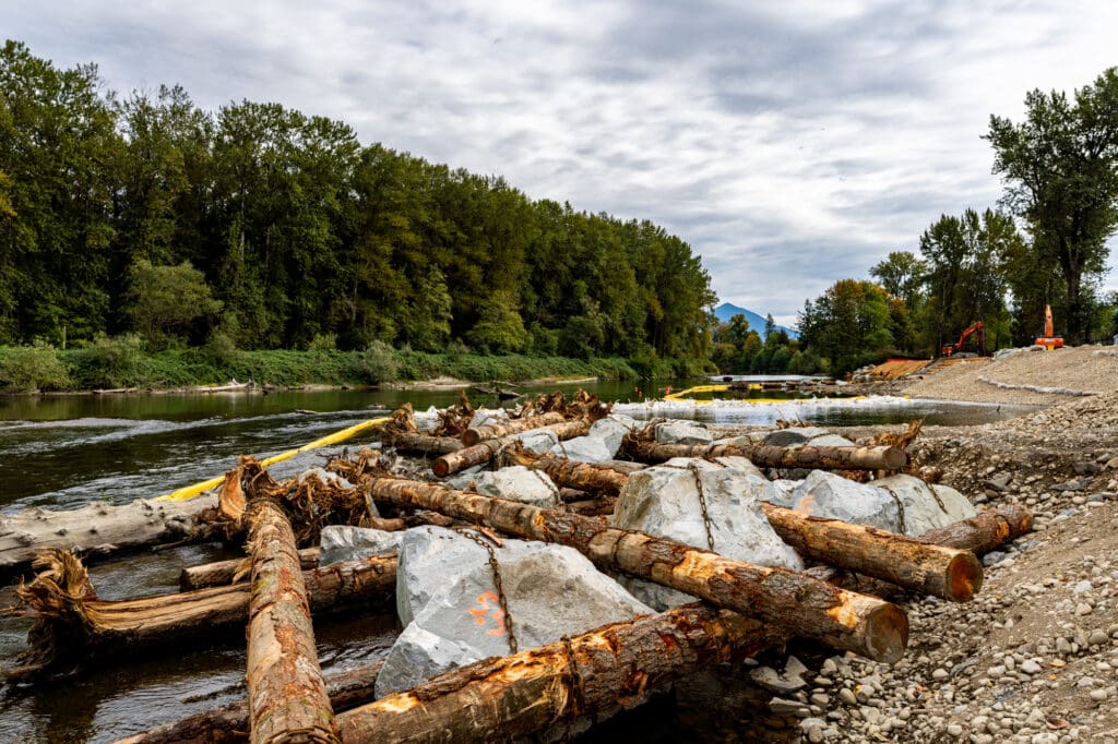 A network of logs anchored to oversized quarry rocks runs along a riverbank.