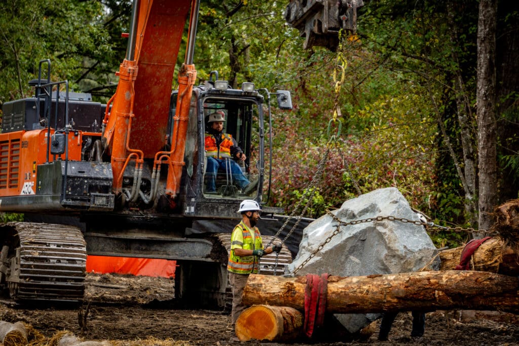 Workers secure the lines on a log-and-rock device while an operator watches from the controls of an excavator.