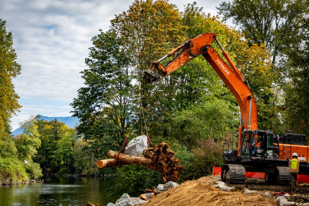 A Hitachi excavator lowers a log-and-rock device down the slope of a riverbank.