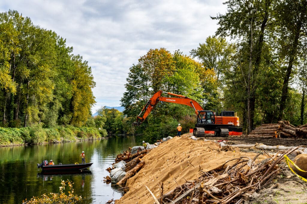 A Hitachi excavator lowers a log-and-rock device onto the base of a riverbank while workers in a fishing boat wait in the river below.