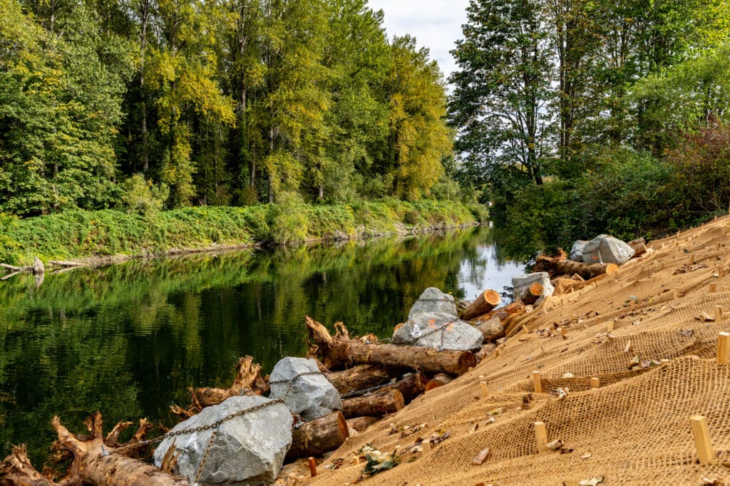 Logs anchored to oversized quarry rocks line the base of a burlap-covered shoreline next to a river.
