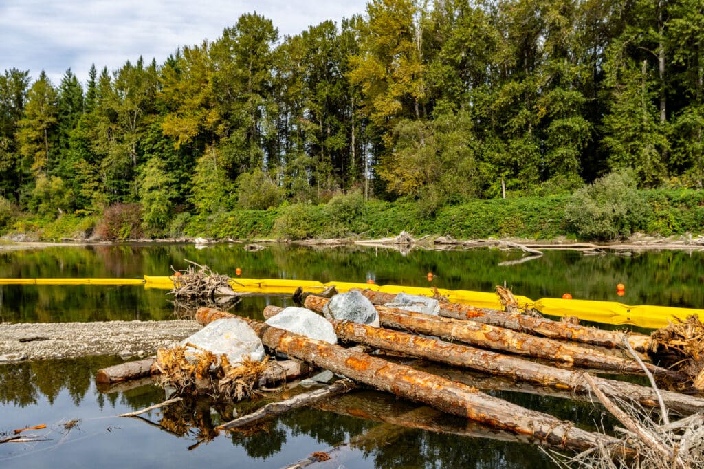 A row of logs is anchored to oversized pieces of quarried rock and rest in the water next to a riverbank.