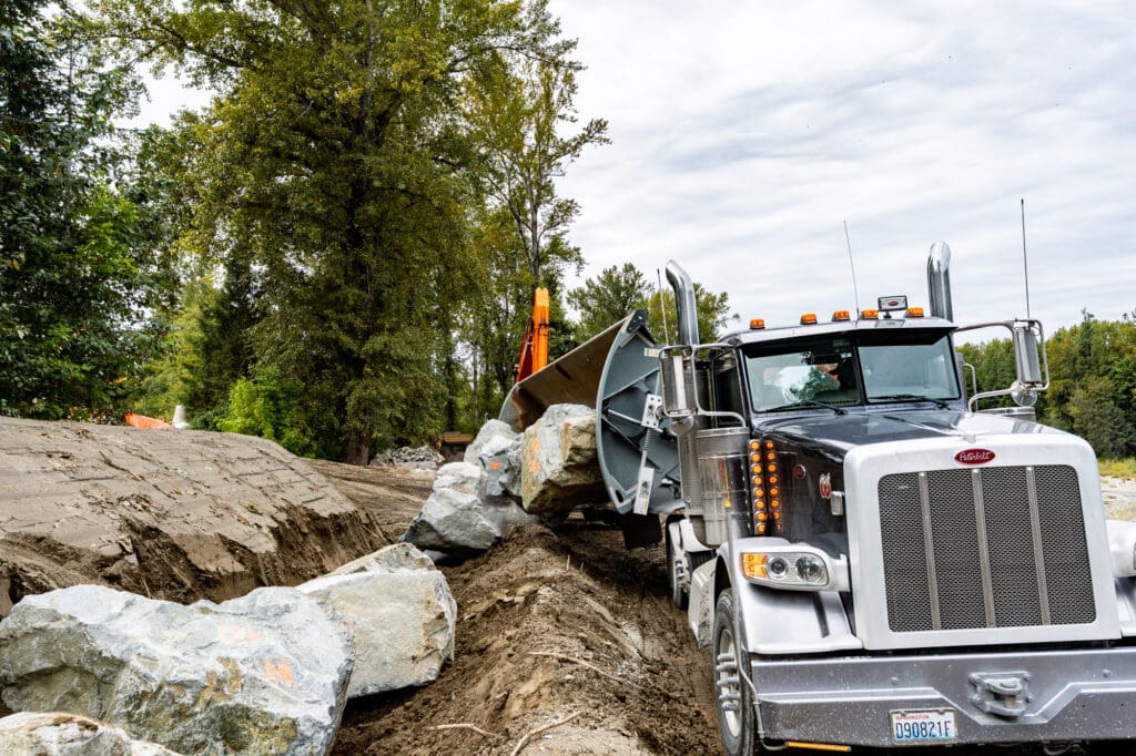 A side-dump truck dumps out a load of 7-Man Quarry Rock into a sandy ditch (preventing breakage).